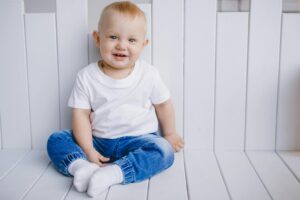 Charming portrait of a smiling baby boy in a white t-shirt and blue jeans, sitting indoors.