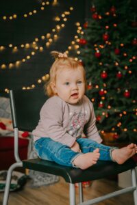 Cute baby in casual clothes sitting on a chair near a festive Christmas tree indoors.