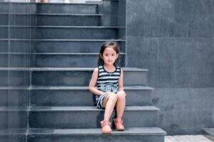 A young girl in a striped dress sits on a sleek grey staircase, looking into the camera.
