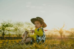 Child in cowboy outfit holding a stuffed toy, sitting outdoors at sunrise.