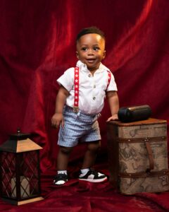 Charming baby boy in red suspenders and shorts posing with vintage decor.