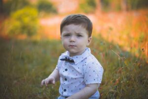 Adorable baby boy sitting outdoors in a sunny park, surrounded by vibrant grass and flowers.