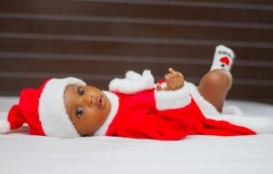 Cute baby in a Santa outfit looking curious, lying indoors on a soft surface.