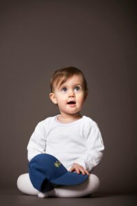 Cute baby sitting indoors, wearing white with a cap, joyful expression.