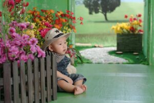 Cute baby sitting by colorful flowers, wearing denim overalls and a cowboy hat.