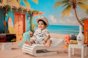 Cute baby dressed in summer attire sitting on a lounge chair in a colorful beach-themed indoor setting.