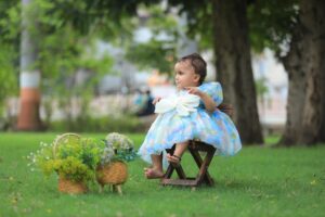 Cute baby girl in a blue dress sitting on a chair in a lush green park, surrounded by trees.