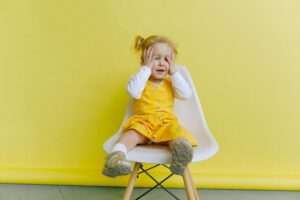 Adorable toddler in bright yellow dress sitting on a chair against a vibrant yellow backdrop, expressing joy.