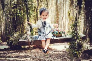 Adorable child smiling on a wooden swing with a basket of flowers in a sunny park.